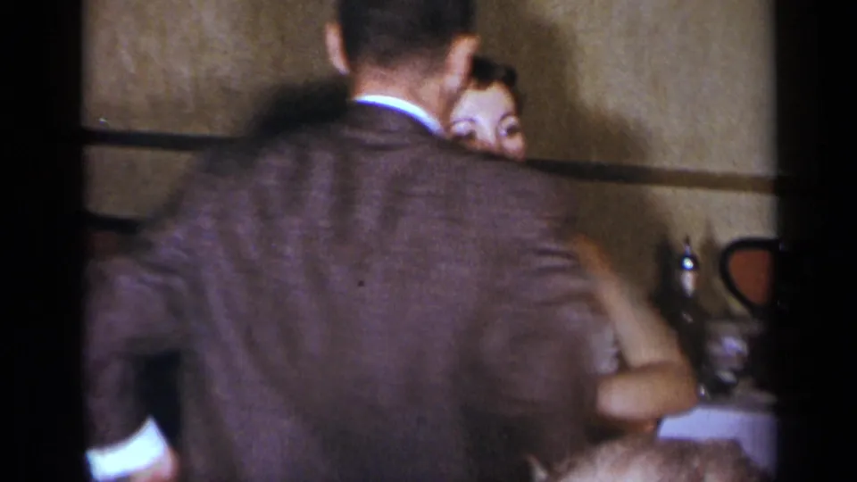 A suited man and woman appear to dance or shift position in a dim 1954 New York restaurant or banquet interior.