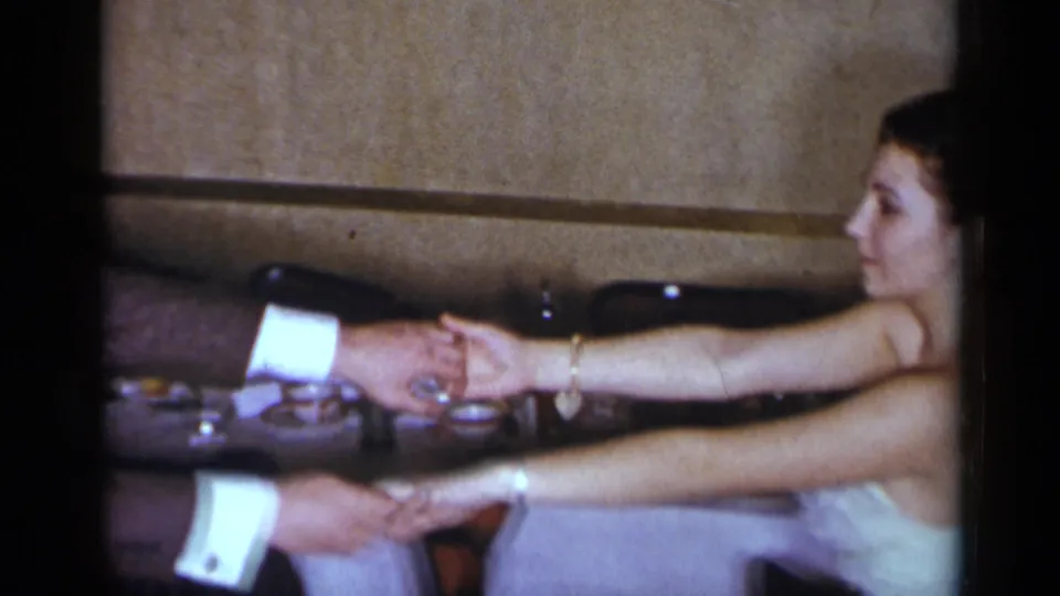 The home-movie frame shows the couple's extended hands and the woman's light dress as they move in a dining-room setting.