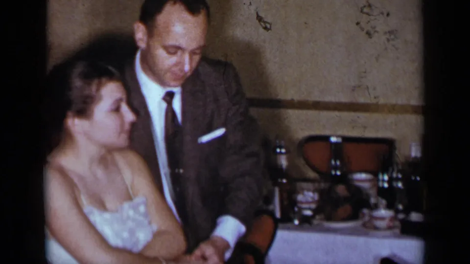 A woman in a light formal dress and a suited man stand close together in a 1954 New York social dining interior.