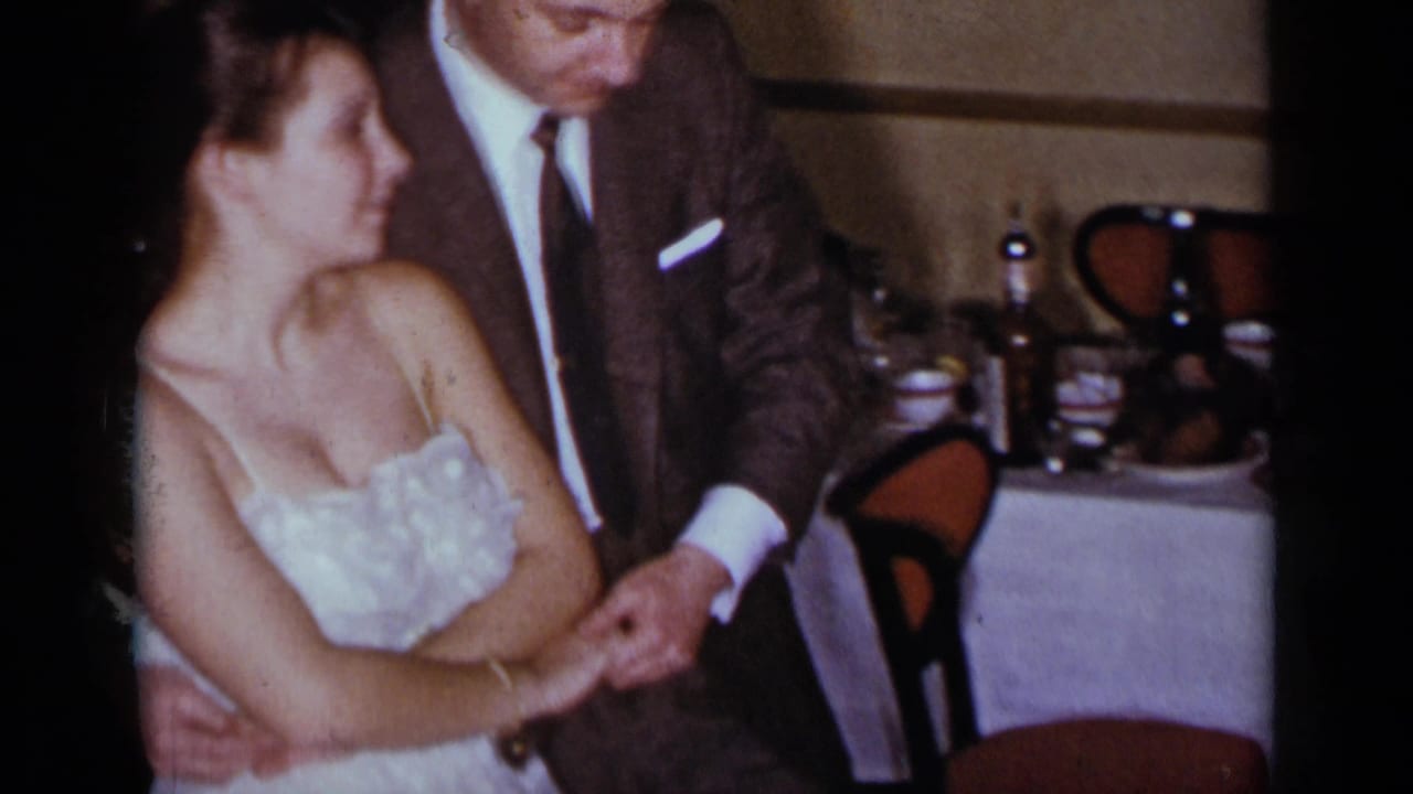 Man in a suit leans over a woman in a white dress while holding her hand.