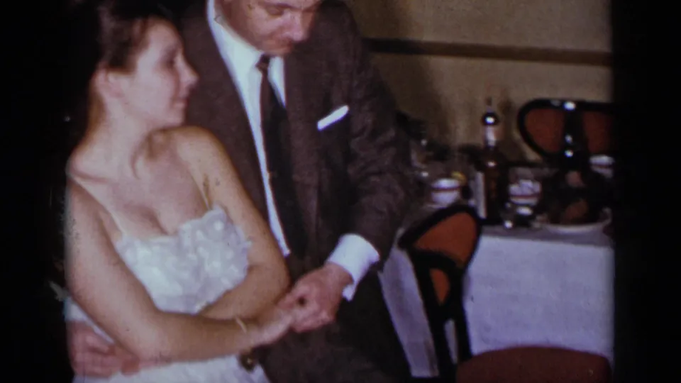 A man in a dark suit leans toward a woman in a white dress as they hold hands in a New York dinner setting.