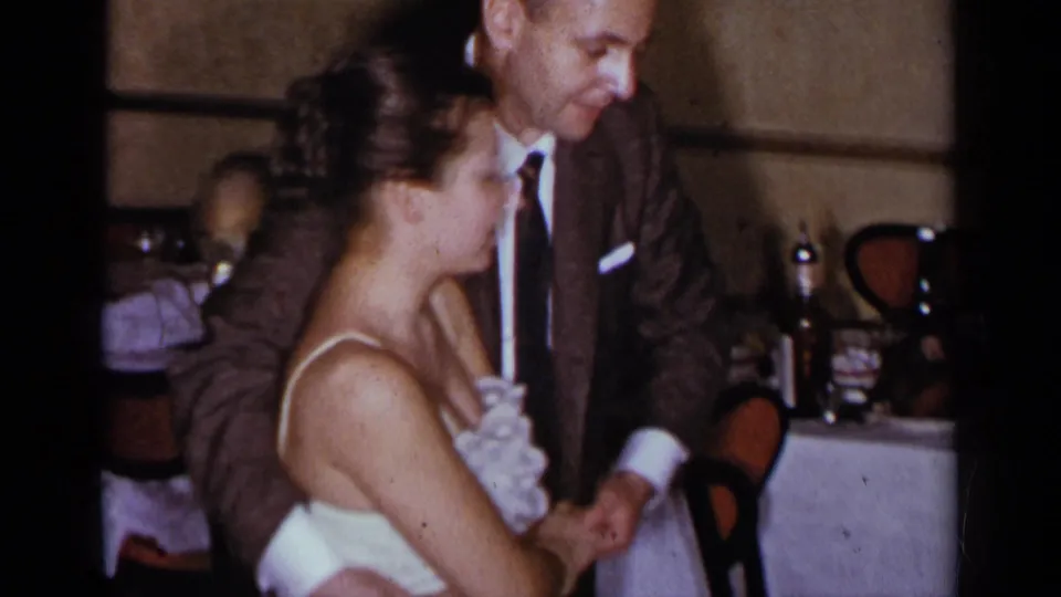 The couple turn toward each other in warm 8mm footage, with table settings and chairs behind them.