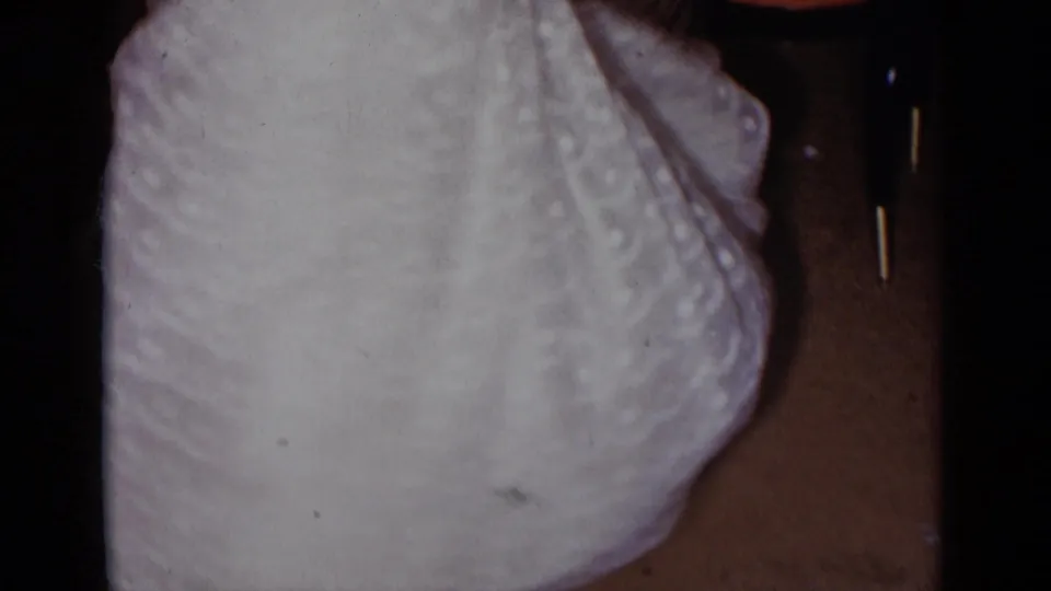 Close cropped 1954 New York home-movie frame showing a white strapless dress in a dim formal interior.