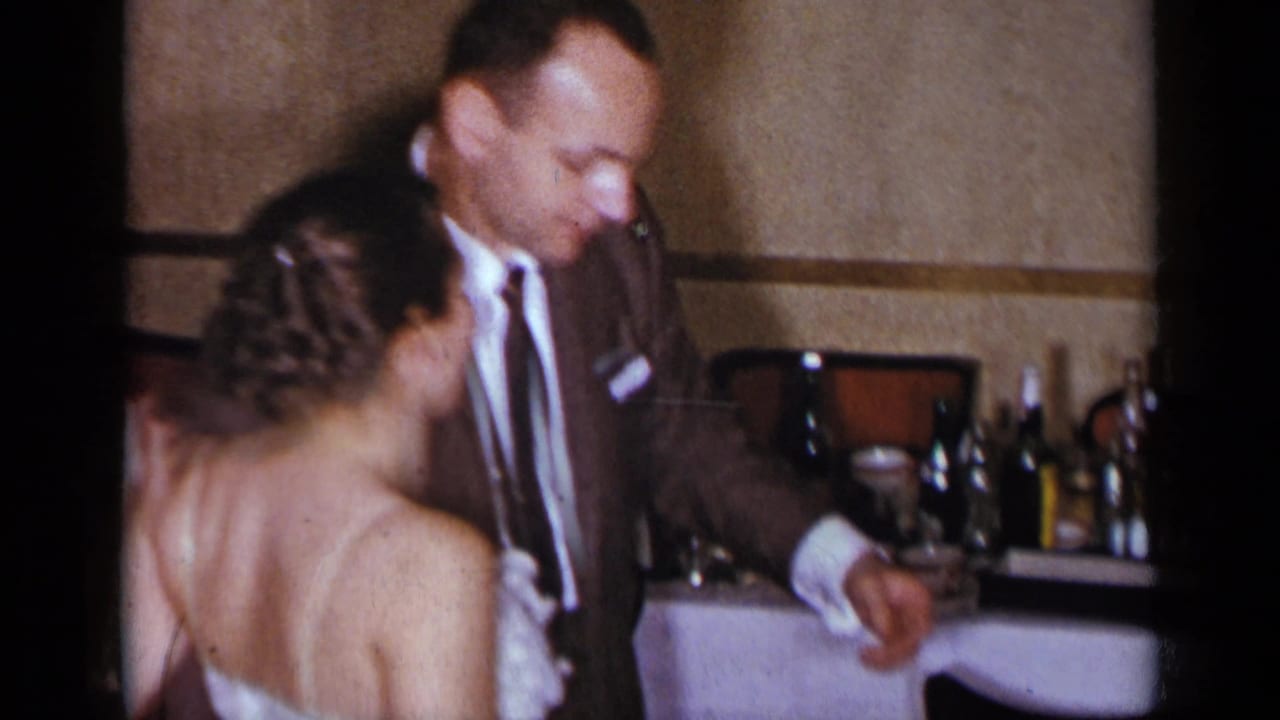 Man in a brown suit dances with a woman in a white dress beside a party table.