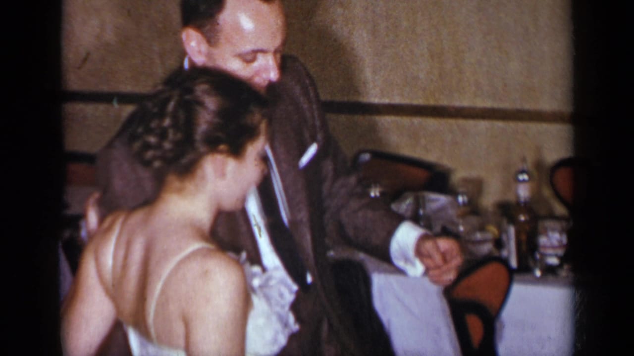 Suited man and woman in white dress dance near a table with bottles and glasses.