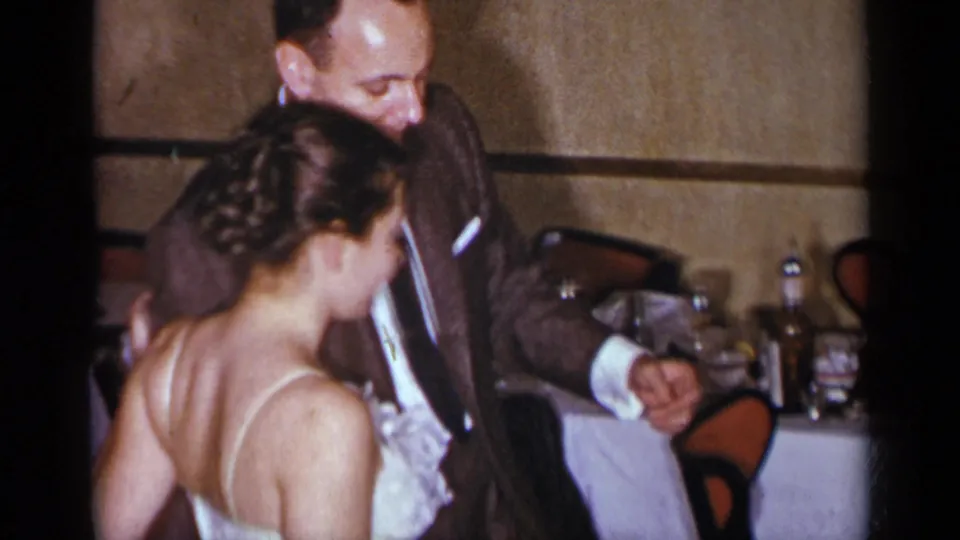 The man gestures while dancing with the woman in a white dress in a warmly lit 1954 New York party interior.
