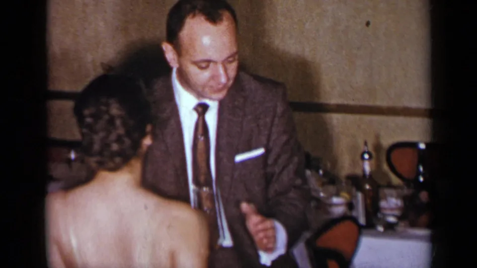 A woman in a white dress faces a suited man as they dance in a mid-century New York party setting with tableware behind them.