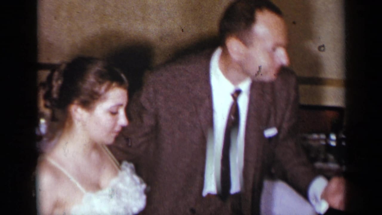 Woman in a white dress stands beside a man in a brown suit in a warm indoor room.