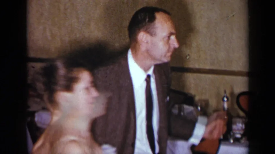 The man in a brown suit and narrow tie turns his head while the woman in a white dress passes in front of him in the indoor New York scene.
