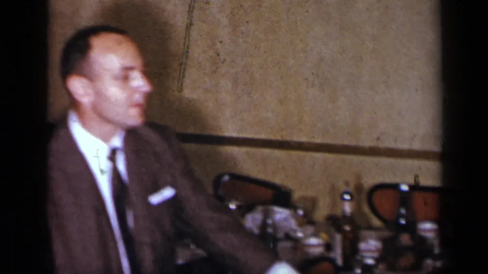 A man in a brown suit and tie appears seated or leaning near dining tables in the warmly lit 1954 New York home-movie interior.