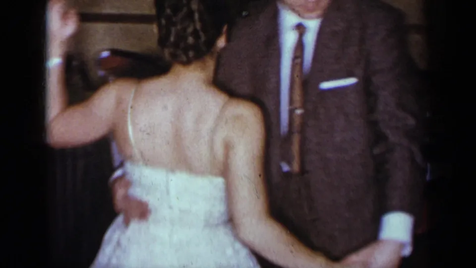 The woman in a white dress is filmed from behind as she dances closely with the man in a brown suit in the indoor New York gathering.