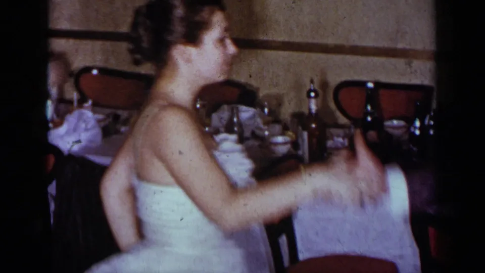 1954 New York 8mm frame of a woman in a strapless white dress turning during a dance, with tables and red-backed chairs behind her.