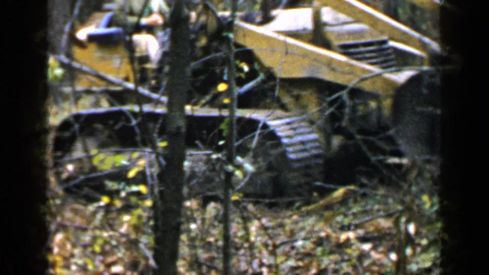 The yellow tracked construction vehicle fills more of the frame as it moves through wooded terrain near Birmingham, Alabama.