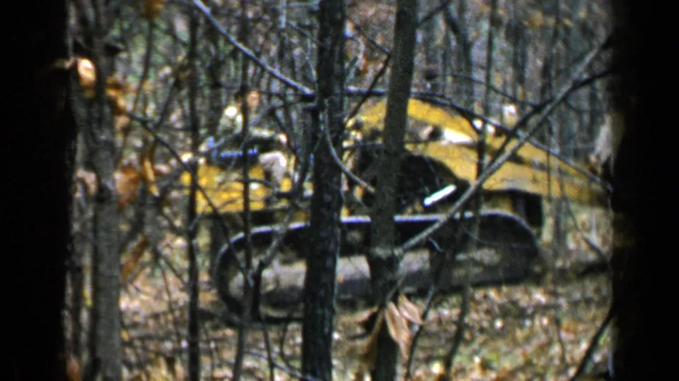 The compact tracked vehicle is partly obscured by dense branches as it continues through the wooded clearing area in 1957 home-movie footage.