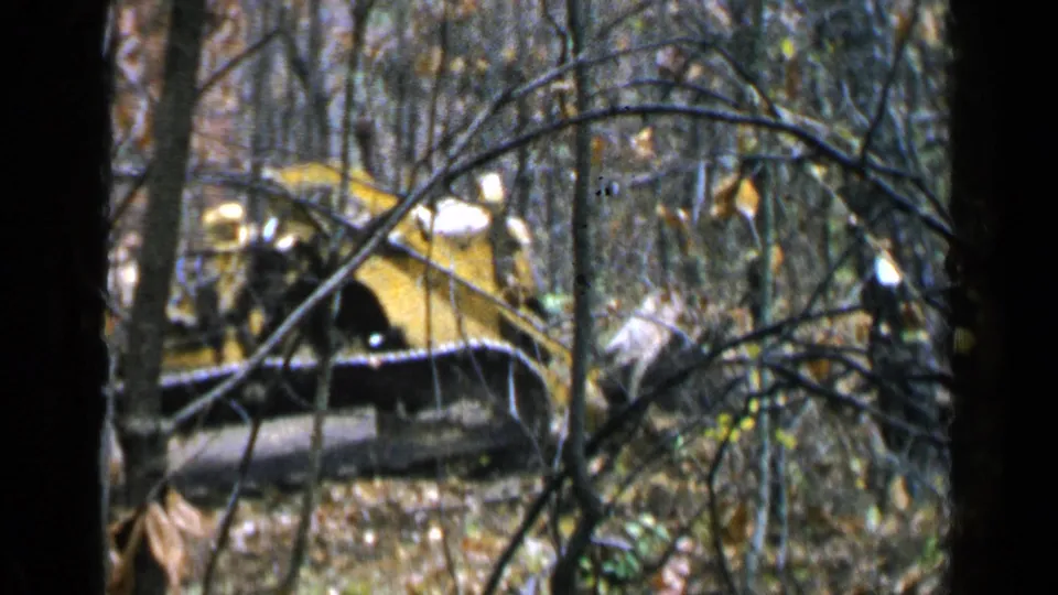 1957 8mm frame of a yellow excavator in a forest, with branches crossing the view and autumn leaves below.