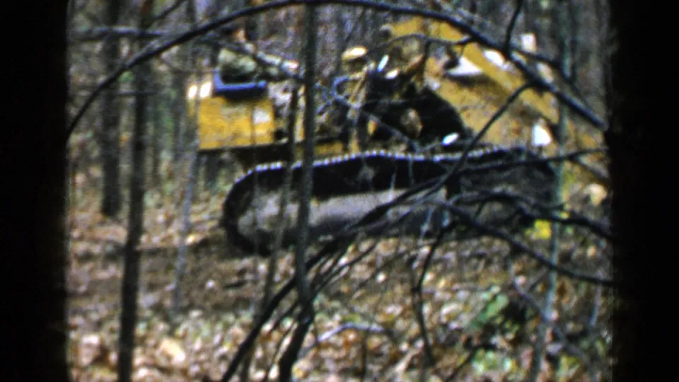 A yellow tracked excavator is partly obscured by trees, with small figures visible on or near the machine in 1957 Alabama footage.