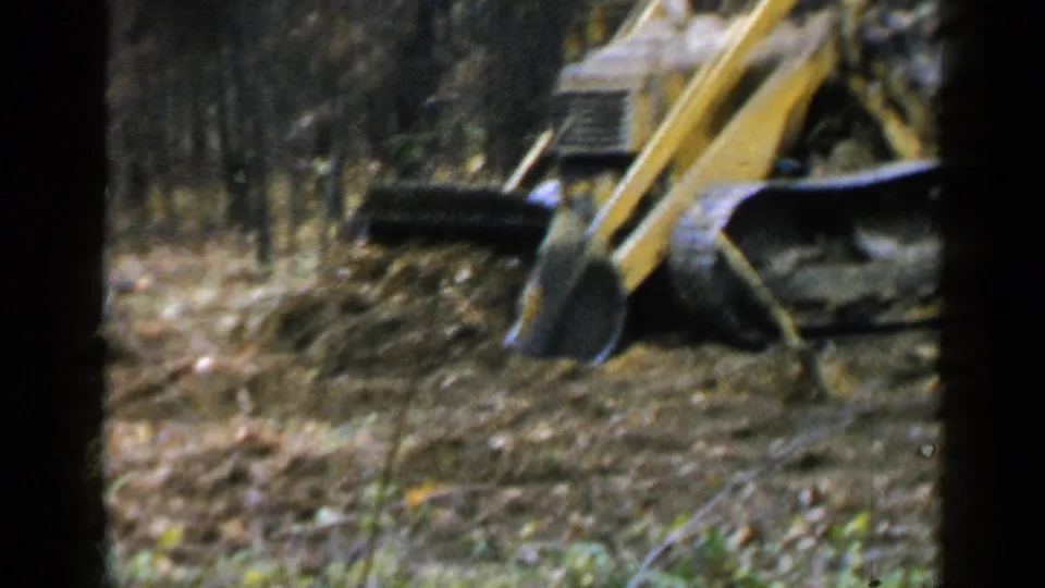 A yellow tracked machine’s arm and bucket work through wooded ground in the 1957 Birmingham-area home-movie footage.