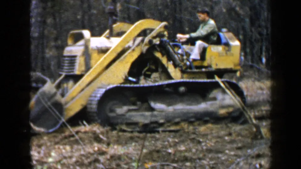 An adult man operates a yellow tracked bulldozer in a wooded Birmingham-area clearing in 1957 home-movie footage.