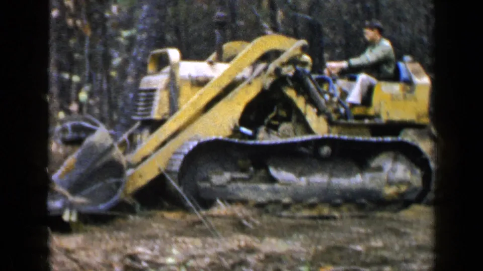 In Birmingham, Alabama, 1957, a yellow tracked bulldozer moves through trees with a man operating it from the seat.