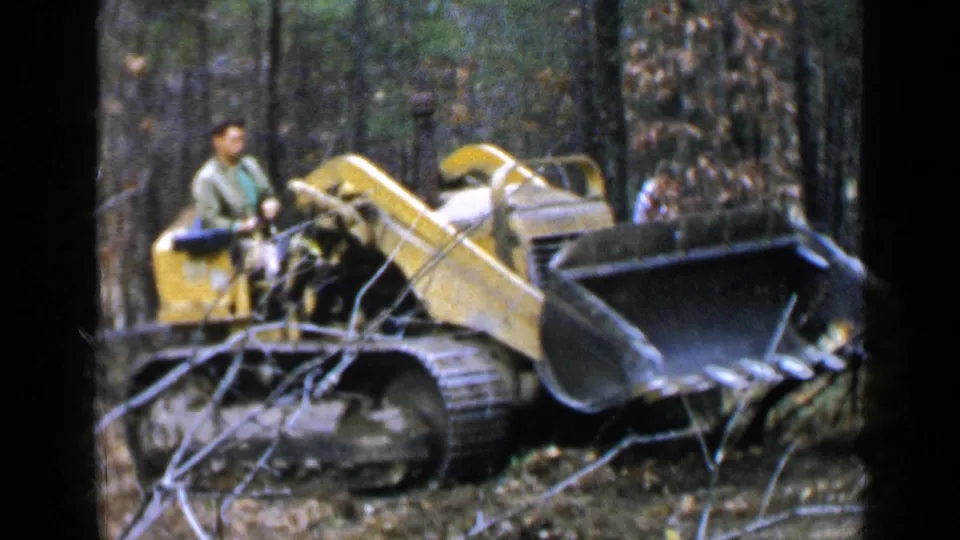 A man drives the yellow tracked loader through a wooded Birmingham path, framed by branches and soft 8mm grain.