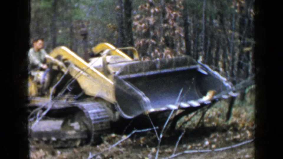 An adult stands beside the yellow front-end loader in wooded Birmingham terrain as the bucket faces forward in the 1957 footage.
