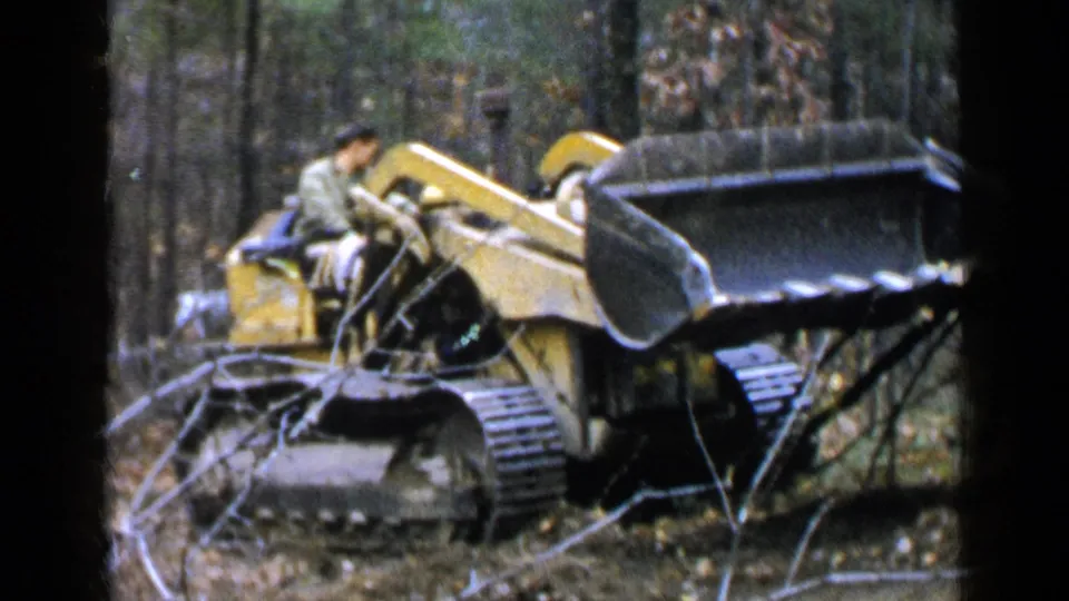 A yellow tracked excavator raises its bucket while a seated operator works among trees and brush in 1957 Birmingham footage.