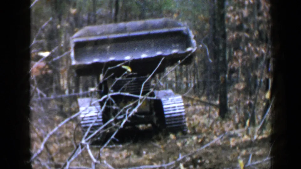 The excavator faces forward with its bucket prominent and the operator partly hidden by trees and brush in the 1957 wooded worksite scene.
