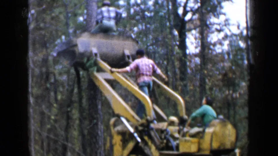 A man in a red plaid shirt climbs onto a raised yellow logging machine in wooded Birmingham, Alabama footage from 1957.