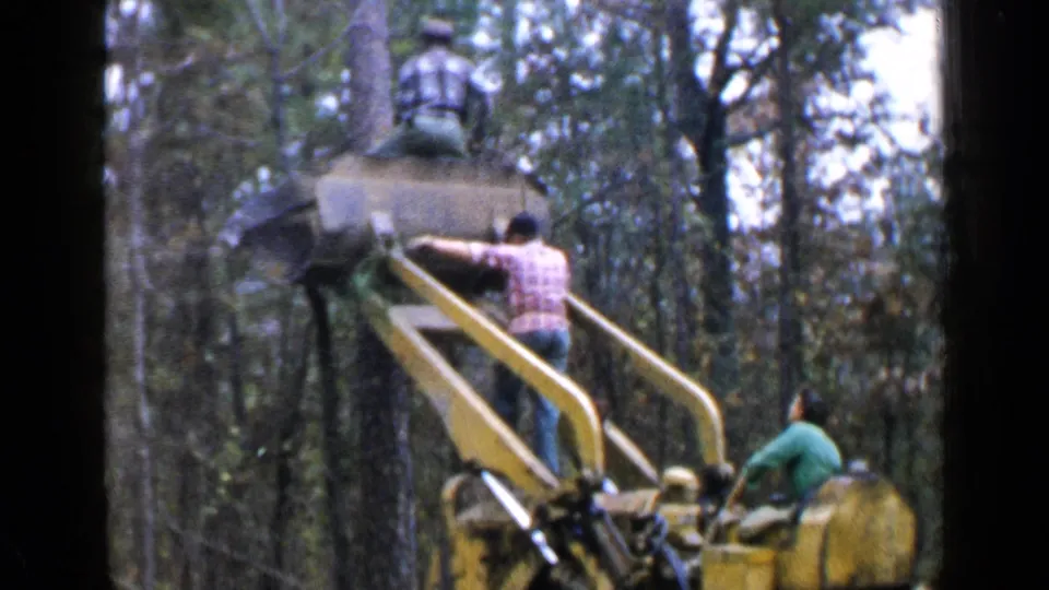 The 1957 Birmingham home-movie frame shows a worker on the ladder of a yellow forestry machine with trees behind him.