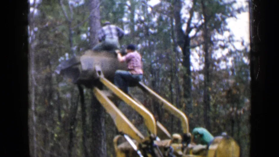 A man in a plaid shirt is near the elevated platform of a yellow logging machine while another worker remains near the cab.