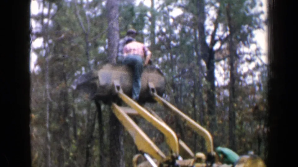 1957 Birmingham-area home-movie frame of a worker operating yellow logging machinery in the woods.