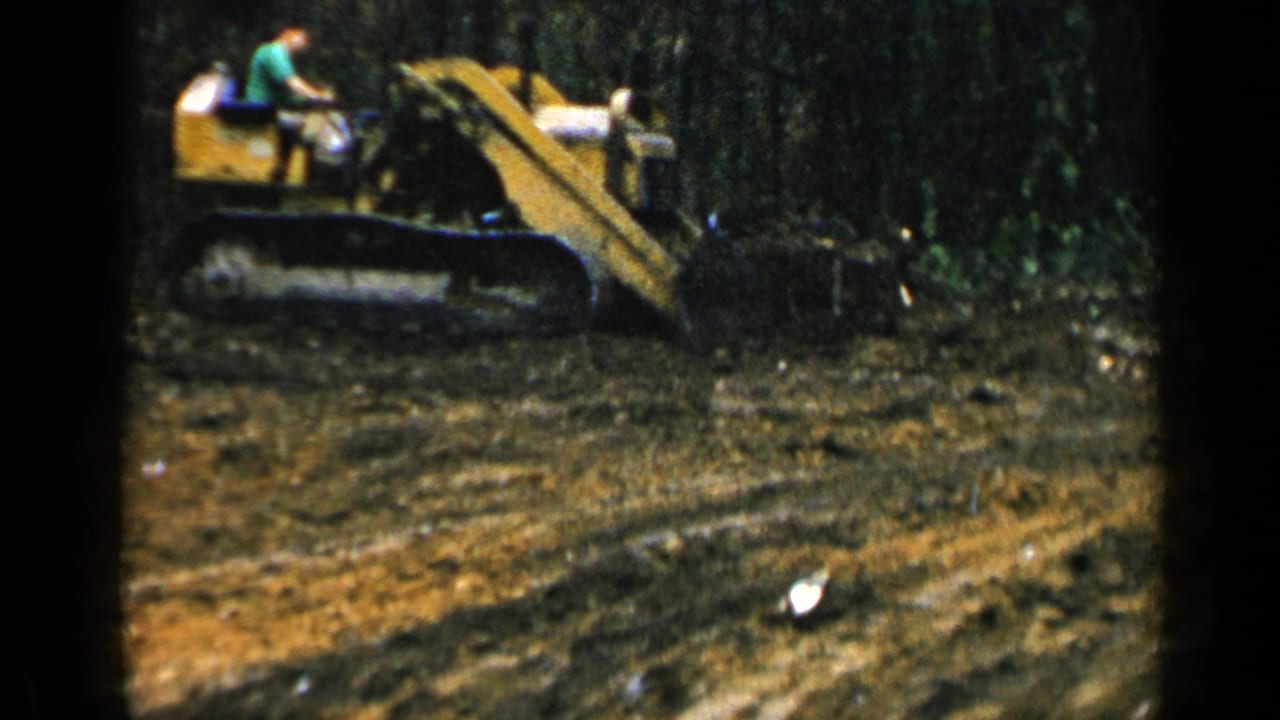 1957 Birmingham 8mm frame showing a man in a green shirt operating a yellow bulldozer beside dense trees and disturbed soil.