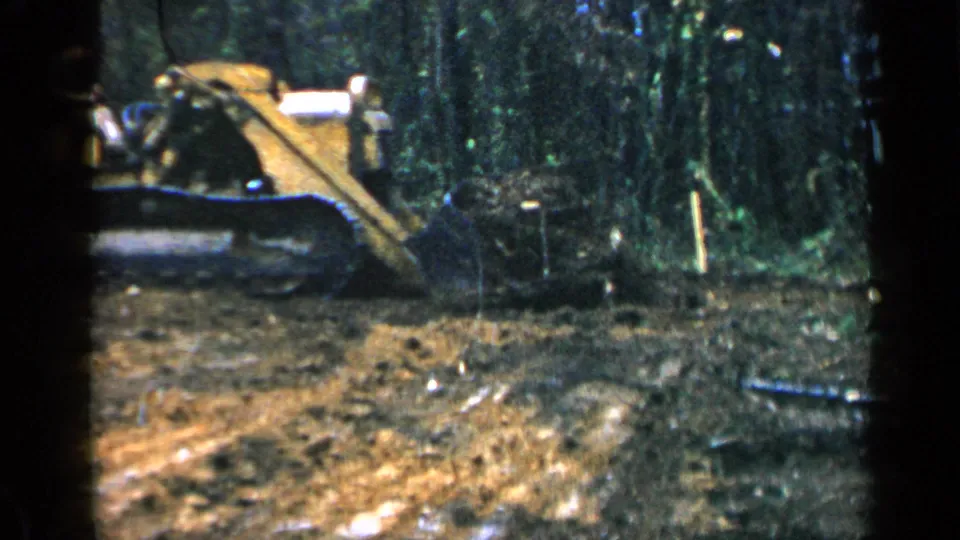 1957 Birmingham home-movie frame of a yellow bulldozer at the edge of dense woods, with disturbed earth in the foreground.