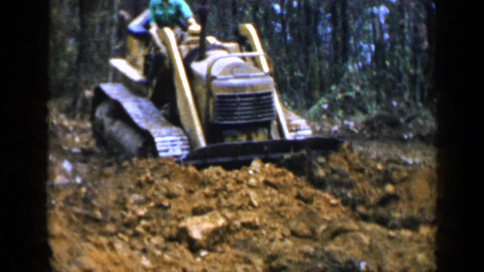 The operator in a green shirt drives a yellow tracked bulldozer through exposed dirt and brush in the Birmingham-area 1957 footage.