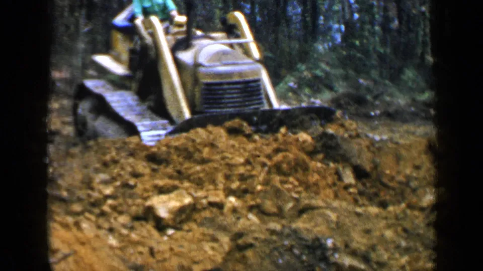 A yellow bulldozer advances through wooded Birmingham terrain, pushing loose soil and rocks in 1957 home-movie footage.