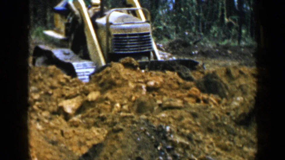 1957 home-movie frame with a yellow bulldozer and partly visible operator working behind a pile of rocky soil in Birmingham woods.