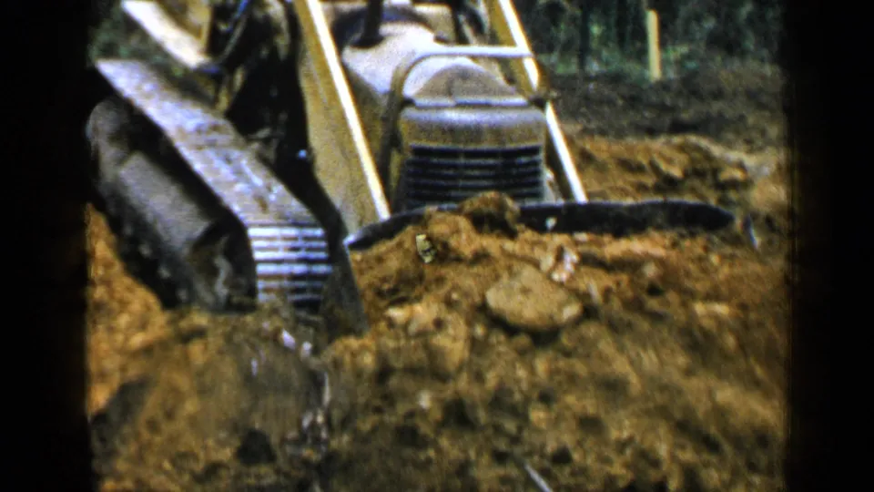 The yellow loader appears in close view as it pushes soil, with woods and rough ground behind it in the 1957 Birmingham footage.