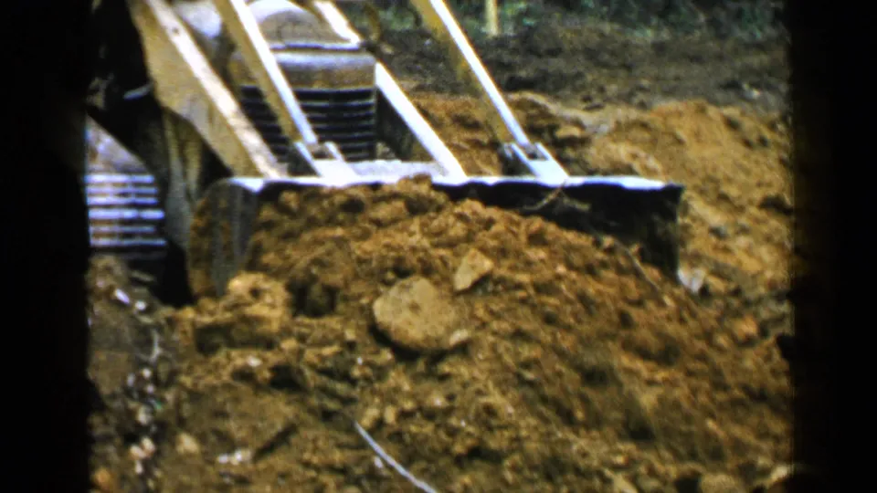 Close 8mm view of the yellow loader's bucket and tracks working through brown soil at a wooded Birmingham-area site.
