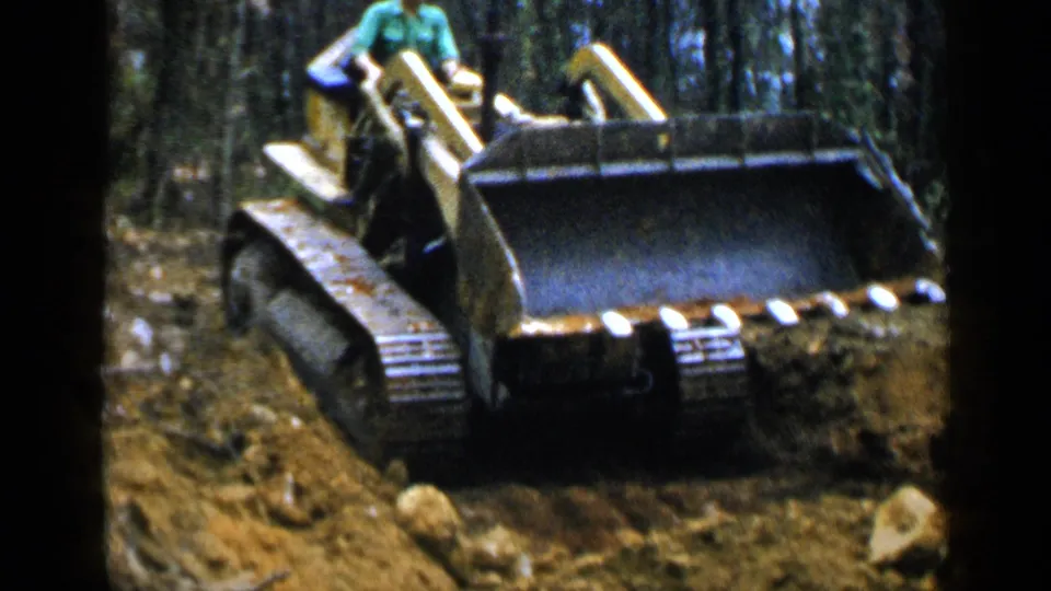 A man in a green shirt operates the yellow tracked loader, its front bucket visible as it moves through the wooded Birmingham worksite.