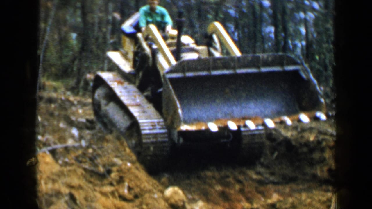 Yellow tracked bulldozer pushes dirt in a wooded area as a man sits at the controls.