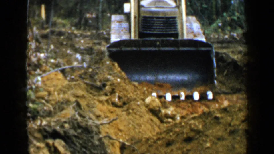 The bulldozer continues forward through wooded ground, its front blade low and carrying soil in the 1957 home-movie scene.