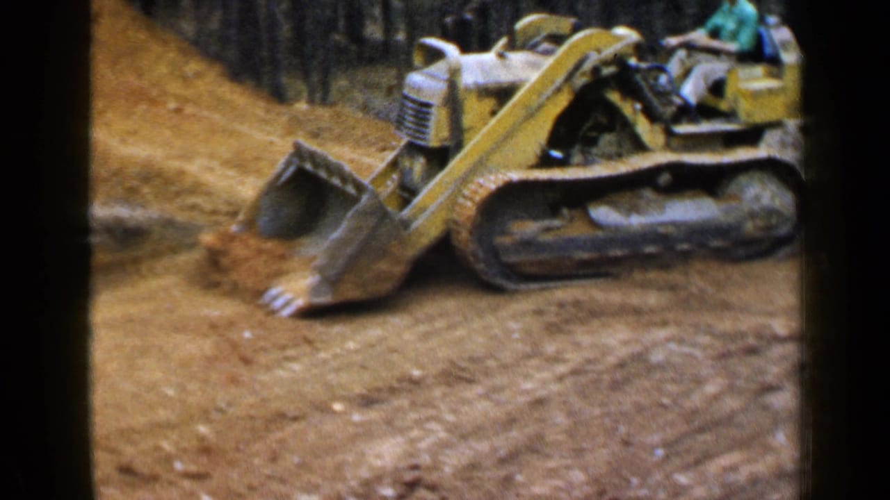 Yellow tracked loader with an operator on a dirt road near woods.