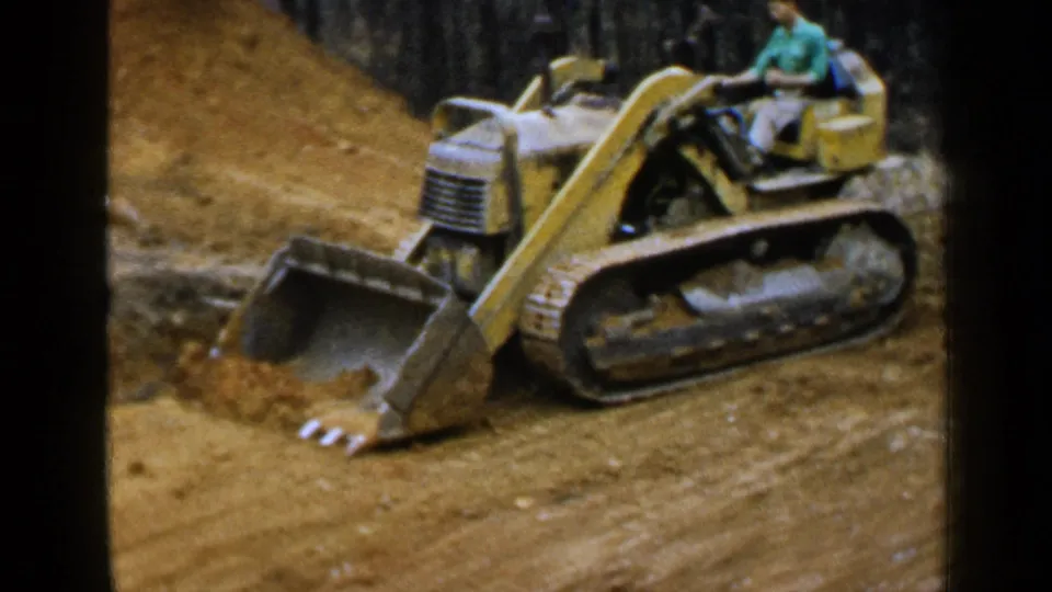 Home-movie frame of a yellow tracked loader and operator moving across reddish dirt near woods in 1957 Alabama footage.