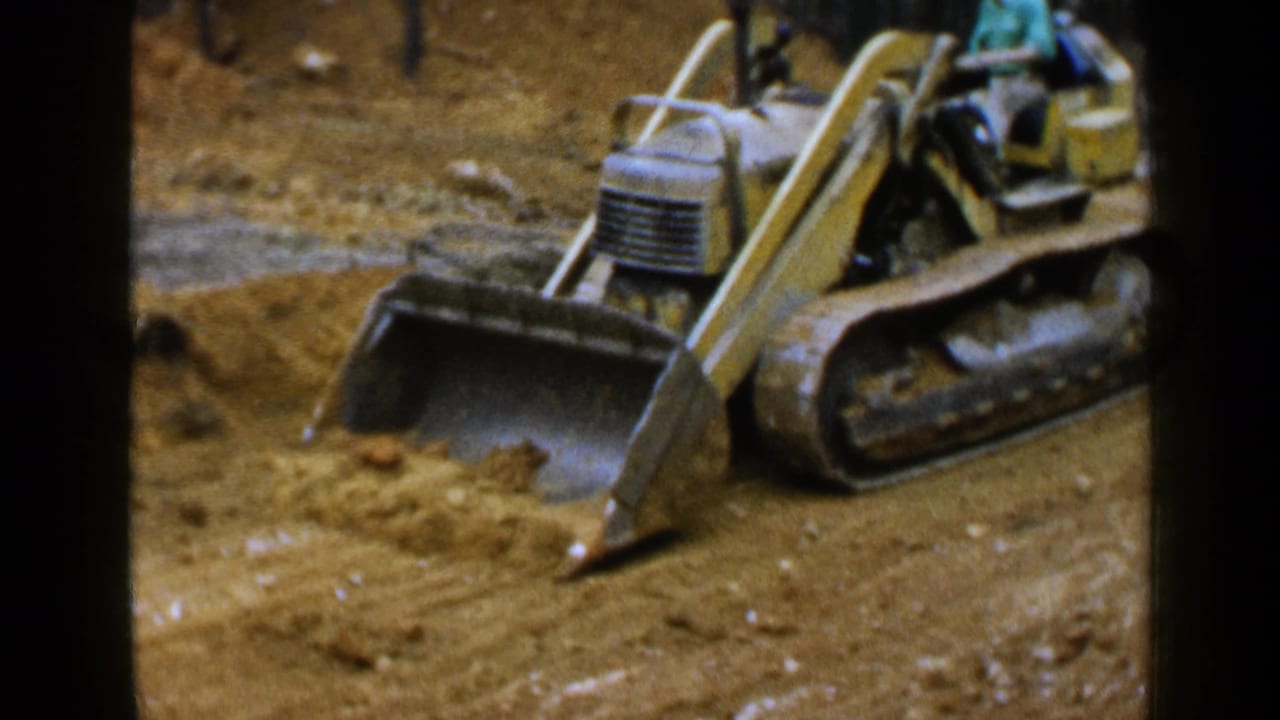 1957 Birmingham home-movie frame showing a yellow tracked loader with a metal bucket moving sand at a construction site.