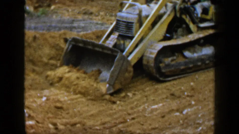 1957 Birmingham frame of a yellow construction loader advancing into sand, with the tracked base and bucket clearly visible.