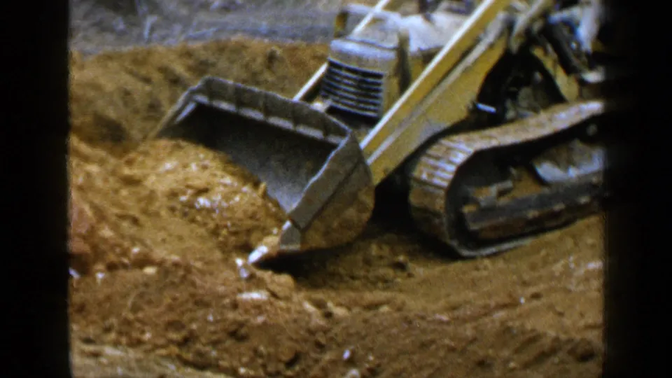 1957 Birmingham 8mm frame showing the loader’s bucket angled into sand, with dark borders and warm film color.