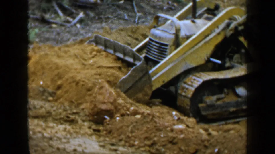 A mid-century tracked loader continues clearing red-brown earth, with its scoop partly buried in the dirt.