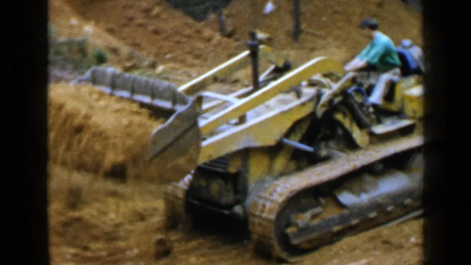 1957 Birmingham frame with an adult operator in a green shirt driving a yellow tracked bulldozer across loose soil.