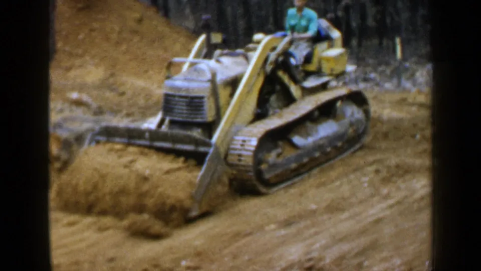 1957 Birmingham frame showing a yellow tracked bulldozer pushing soil forward, with an adult operator seated above the treads.