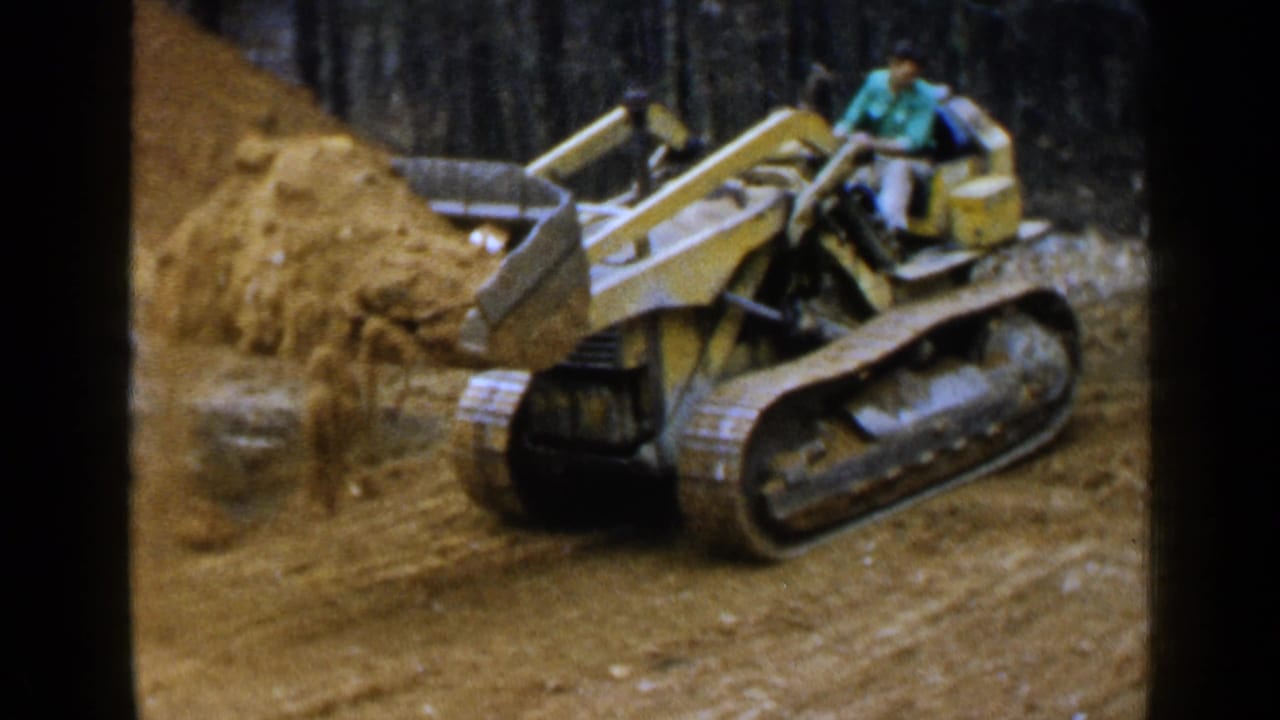 Man operating a yellow tracked loader beside a mound of dirt in a wooded worksite.